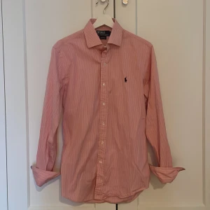Rosa randig skjorta Polo Ralph Lauren - Snygg rosa skjorta från Polo Ralph Lauren i slim fit med tunna vita ränder och klassisk krage. Broderad logga på bröstet och knäppning framtill. Långärmad modell i bomull som passar perfekt till jeans eller chinos.