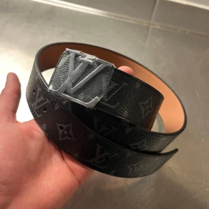  svart monogram bälte - Snyggt svart bälte  med klassiskt monogrammönster och stor spänne i silver. Bältet är gjort i skinn med en lyxig känsla och har en insida i ljusbrunt. Perfekt accessoar för att lyfta din outfit.