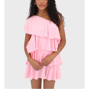 Rosa volangklänning från Meet Me There - Supersöt rosa kortklänning med flera volanger och oneshoulder-design. Klänningen har en luftig och flowy känsla, perfekt för sommarens alla tillfällen. OBS den är kort i modellen. Säljes med prislapp kvar och är oanvänd 
