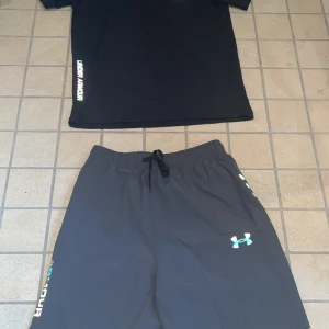 Svart Under Armour t-shirt med gråa shorts - Svart t-shirt från Under Armour med diskret logga och text längs sidan. Tillverkad i ett lätt och svalt syntetmaterial, perfekt för träning. Klassisk rund hals och korta ärmar. Snygg och sportig stil som funkar till alla aktiva dagar. Även gråa shorts till.