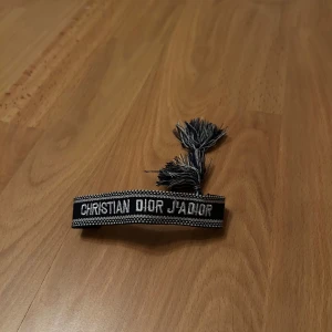 Christian Dior J'Adior armband - Snyggt svart och vitt armband från Christian Dior med texten 'J'Adior' broderad framtill. Armbandet har två coola tofsar i ändarna och är gjort i ett vävt tygmaterial. Perfekt accessoar för att lyfta din outfit med en lyxig touch.
