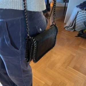 Svart axelväska med kedjedetaljer - Svart mini axelväska! Väskan har silver kedja, det finns en ”ficka” som är liten inuti där man t.e.x kan lägga sitt kort i eller så!