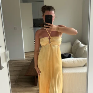 Ljusgul plisserad långklänning - Superfin ljusgul långklänning med plisserad kjol och snygga smala axelband som korsas i ryggen. Klänningen har öppen rygg och knyt i midjan, samt en liten cut-out framtill. Perfekt för sommarens alla tillfällen!