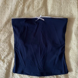 Mörkblå bandeau tubtopp med rosett XS - Supersöt mörkblå bandeau tubtopp med vit spetskant upptill och en liten vit rosett i mitten. Toppen är ärmlös och har en enkel, tight passform. Perfekt att styla med jeans eller kjol för en trendig look.