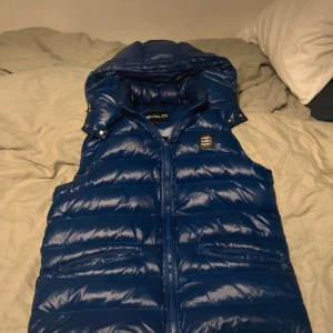 Blå dunväst från Bvaldi med huva - Snygg blå dunväst från Bvaldi med glansig finish och avtagbar huva. Västen har dragkedja framtill, två fickor med dragkedja och ett svart logomärke på bröstet. Perfekt för lager-på-lager och chillare dagar. Har en liten difekt på nackdelen men sys knappt. Nypris 1899kr