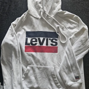 Vit hoodie från Levi's med logga stl XS - Säljer en vit hoodie från Levi's i storlek XS. Tröjan har stor tryckt logga i blått, vitt och rött framtill, huva med snörning och känguruficka. Mjuk bomullskänsla och ribbade muddar. Perfekt för en chill och stilren look.