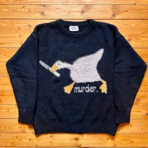 Svart stickad tröja med gås - Svart stickad tröja från Cuang yi med ett stort motiv av en gås som håller en kniv och texten 'murder.' på framsidan. Tröjan har rund hals, långa ärmar och ribbade muddar. Perfekt för dig som gillar humoristiska och unika plagg.