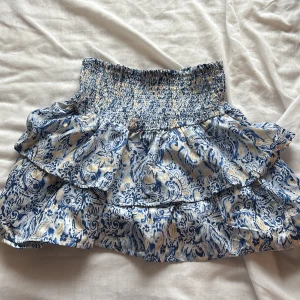 Blåvit volangkjol med smock från Gina tricot - Supersöt kort kjol med volanger och smockad midja. Mönstret är blått och beige på vit botten, vilket ger en fräsch och somrig vibe. Kjolen är luftig och lätt, perfekt för varma dagar. Passformen är normal och materialet känns som tunn bomull eller viskos.