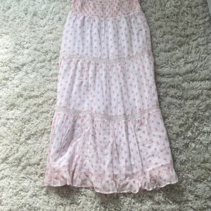 Vit maxi/långkjol med rosa blommor - Bara använd en gång och har bara dragit av lappen. Supersöt maxikjol i vitt med små rosa blommor, volangkant nertill och smock upptill. Kjolen har spetsdetaljer och är gjord i ett lätt bomullstyg, perfekt för varma dagar. Den har en romantisk och bohemisk vibe som passar perfekt till sommaren.