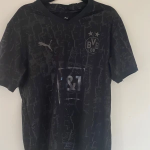 Svart BVB Puma fotbollströja S - Snygg svart Borussia Dortmund fotbollströja från Puma med diskret mönster och silvriga detaljer. Tröjan har korta ärmar, BVB-logga på bröstet och sponsortryck i silver. Tillverkad i lätt och funktionellt material som passar perfekt för träning eller match.