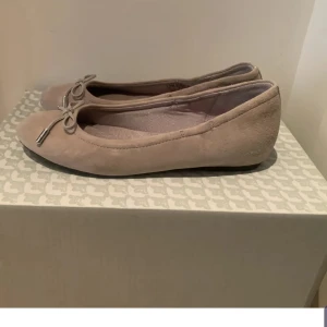 Beige ballerinaskor med rosett - Snygga beige ballerinaskor i mocka med rund tå och dekorativ rosett framtill. Skorna har platt sula och en stilren, klassisk design som passar till många outfits. Perfekta för dig som gillar en enkel och chic look.