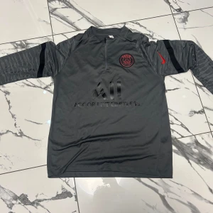 PSG långärmad träningströja Nike M - Snygg grå långärmad fotbollströja från Nike för Paris Saint-Germain. Tröjan har svart PSG-logga och röd Nike-swoosh på bröstet, samt diskret mönstrade ärmar. Materialet är lätt och andas, perfekt för träning eller match.