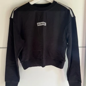 Svart Calvin Klein sweatshirt - Svart sweatshirt från Calvin Klein Performance med vita detaljer på axlarna och logga framtill. Tröjan har en klassisk rund halsringning och långa ärmar. Perfekt för en sportig och stilren look.