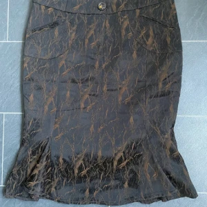 Svart-brun kjol  med mönster - Unik kjol med brunt abstrakt mönster. Kjolen har fickor fram, dekorativa knappar och plisserad nederdel bak. Snygg midjemodell med coola detaljer och knappstängning. Perfekt för dig som gillar statement-plagg.
