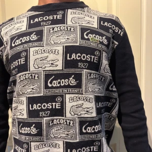 Mönstrad stickad tröja från Lacoste - Snygg stickad tröja från Lacoste i marinblått och vitt med coolt all-over logomönster och klassisk rund hals. Tröjan har långärmade ärmar och är perfekt för dig som gillar streetstyle och vill sticka ut med en ikonisk look. Köpt i Frankrike förra året