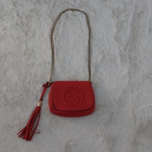 Röd axelväska från Gucci i skinn - Säljer en snygg röd axelväska från Gucci i äkta skinn med guldfärgad kedja och stor tofsdetalj. Väskan har präglad GG-logga på framsidan och ett ljust innerfoder med ficka. Perfekt storlek för det viktigaste och ger en lyxig touch till din outfit.