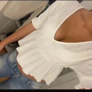 Vit v-ringad blus från Pull&Bear - Supersöt vit blus från Pull&Bear med v-ringning och korta ärmar. Modellen är croppad med volangkant nedtill och lätt rynk vid midjan. Perfekt för dig som gillar en luftig och feminin stil. Tunt, strukturerat tyg som känns skönt mot huden.