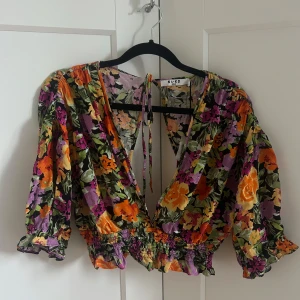 Blommig omlottblus från NA-KD - Färgstark blommig omlottblus från NA-KD med lila, orange och gröna toner. Djup v-ringning, elastisk midja och trekvartsärmar med volangdetaljer. Öppen rygg med knytband. Aldrig använd då jag insåg för sent att det inte var mina färger. 