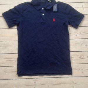 Marinblå pikétröja från Ralph Lauren - Snygg marinblå pikétröja från Polo Ralph Lauren med klassisk krage, korta ärmar och två knappar framtill. Ikoniska röda loggan broderad på bröstet. Tillverkad i bomull för en skön och stilren look.