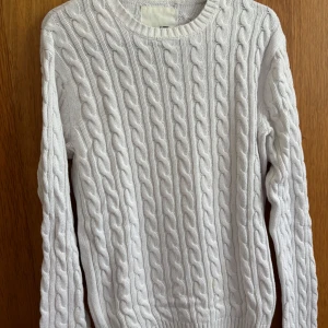 Vit kabelstickad tröja i bomull - Kabelstickad vit tröja med rund hals och långa ärmar. Tröjan har klassiskt flätmönster och är gjord i mjuk bomull, perfekt för lager-på-lager under kyliga dagar. Snygg och enkel att matcha med jeans eller kjol.
