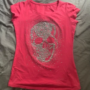 Röd t-shirt med döskalle i pärlor - Cool röd T-shirt med korta ärmar och rund halsringning. Framsidan har en stor döskalle dekorerad med silvriga pärlor som ger en edgy vibe. Perfekt för dig som vill sticka ut med en unik detalj. Materialet känns mjukt och skönt mot huden. Den har tappat några av diamanterna men inget man tänker på!