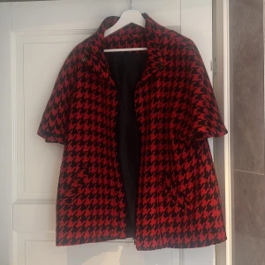 Röd och svart rutig kappa med kort ärm - Snygg kappa i röd och svart hundtandsmönster med korta ärmar och bred krage. Jackan har två fickor framtill och stängs med knappar upptill. Perfekt för dig som vill sticka ut med ett klassiskt men ändå edgy plagg.