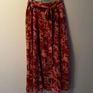 Röd blommig långkjol från Indiska - Säljer en superfin långkjol från Indiska i rött och rosa med blommigt mönster. Kjolen har en bekväm resår i midjan och ett snyggt knytband framtill. Katt finns i hemmet, kontakta vid fler frågor🌷