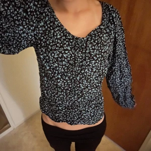 Svart blommig blus med puffärm - Supersöt svart blus med småblommigt blått mönster. Blusen har långa puffärmar och en lätt croppad passform. Tyget är tunt och flowy, perfekt för vår och sommar. Snygg v-ringning och smockad nederkant som ger en fin siluett.