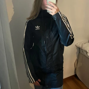 Regnjacka adidas - Supersnygg regnjacka från Adidas. Jackan har huva, dragkedja och knappar framtill samt snörning i halsen. Använd fåtal gånger 💕💕