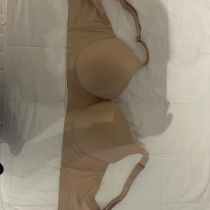Beige bh 95E - Säljer en beige bh med slät form och lätt vadderade kupor. Toppen har justerbara och avtagbara axelband, perfekt under tighta plagg. Materialet är mjukt och stretchigt med 90% polyamid och 10% elastan.