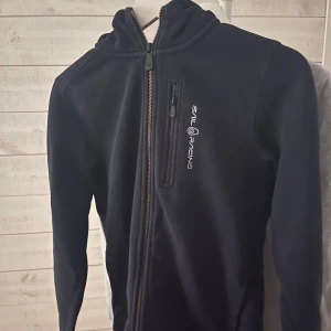 Svart fleecejacka från Sail Racing - Svart ziphoodie från Sail Racing med huva och hel dragkedja framtill. Tröjan har en bröstficka med dragkedja och broderad logga på bröstet. Perfekt för lager-på-lager och chill dagar. Snygg och enkel design som funkar till det mesta. Skriv ifall ni har frågor.