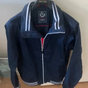Mörkblå vindjacka från Etirel S - Snygg mörkblå vindjacka från Etirel med vita ränder på krage och muddar. Jackan har dragkedja framtill, broderad logga på ärmen och röda detaljer på insidan. Perfekt för dig som gillar sportig och klassisk stil.