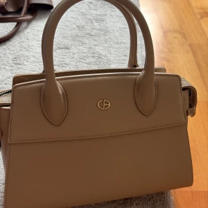 Beige handväska från Chrisbella - Stilren beige handväska från Chrisbella med matchande plånbok. Väskan har två rejäla handtag, avtagbar axelrem och guldfärgade detaljer. Tillverkad i syntetmaterial med slät finish och dragkedja upptill. Perfekt för dig som gillar klassisk och clean look.