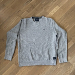 Ljusgrå stickad tröja från Bondelid - Snygg ljusgrå stickad tröja från Bondelid med diskret strukturmönster och broderad logga på bröstet. Klassisk rund halsringning med ribbade muddar vid ärmslut och nederkant. Perfekt för dig som gillar stilrena och bekväma plagg.