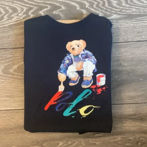 Mörkblå Polo Bear t-shirt från Ralph Lauren - Snygg mörkblå t-shirt från Polo Ralph Lauren med ikoniska Polo Bear-trycket framtill. Björnen målar färgglada bokstäver och har cool outfit. T-shirten är i mjuk bomull och har klassisk passform med korta ärmar och rund hals.