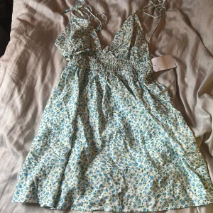 Blommig kortklänning från Zara XS - Supersöt kort klänning från Zara i storlek XS med ljusblått blommönster på vit botten. Klänningen har smock i midjan, tunna knytband och v-ringad hals. Perfekt för varma dagar och ger en fräsch sommarkänsla.