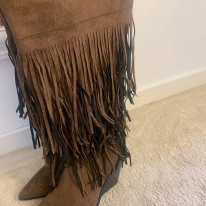 Bruna mocka cowboy boots med fransar - Snygga bruna cowboy boots i mocka med långa fransar längs skaftet och spetsig tå. Perfekt till dig som ska på festival till sommar eller älskar western stil!