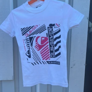 Vit Quiksilver t-shirt med grafiskt tryck - Vit t-shirt från Quiksilver med coolt grafiskt tryck i svart och rött framtill. Klassisk rund hals och korta ärmar. Skön passform och tillverkad i mjuk bomull, perfekt för en avslappnad stil.