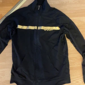 Svart kofta från BOSS med gul detalj - Svart kofta från BOSS med gul rand och logga över bröstet. Koftan har hel dragkedja, hög krage och två fickor framtill. Perfekt för dig som gillar sportig och clean stil med en touch av färg.