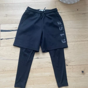 Svarta leggings med shorts från Zara - Svarta leggings med fastsydda shorts från Zara. Byxorna har elastisk midja med snörning och tryckt text på ena benet. Perfekta för träning eller chill, tillverkade i mjukt syntetmaterial för maximal komfort.