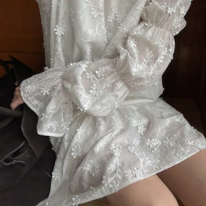 Vit blommig kortklänning med volang - Superfin vit kortklänning med broderade blommor, volang nertill och långa puffärmar. Klänningen har öppen rygg med knytband och är gjord i ett lätt, luftigt material. Perfekt för sommarens alla tillfällen och ger en romantisk vibe.