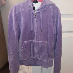 Lila hoodie från Juicy Couture XS - Supermjuk lila hoodie från Juicy Couture i velour-look med dragkedja, huva och snörning. Har broderad JC-logga på bröstet och stora fickor framtill. Materialet är 80% bomull och 20% polyester. Perfekt för en chill och trendig vibe.
