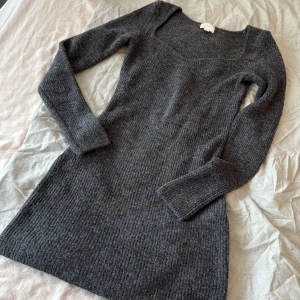 Mörkgrå stickad klänning - Mysig mörkgrå stickad klänning med lång ärm och v-ringad hals. Perfekt för kyliga dagar och enkel att styla med boots eller sneakers. Ribbad struktur ger en snygg och bekväm passform.