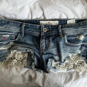 Jeansshorts med spets från Hollister - Snygga blå jeansshorts från Hollister med slitna detaljer och råa kanter. Shortsen har vit spets längs bensluten som ger en extra touch. Klassisk femficksmodell med logga på fickan och knapp framtill. Perfekta för sommaren!