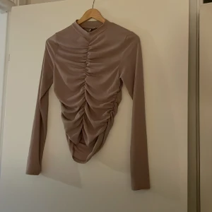Beige ruched topp från NLY Trend XS - Säljer en beige långärmad topp från NLY Trend i storlek XS. Toppen har hög hals och snygg rynkad detalj framtill som ger en cool och trendig look. Materialet är mjukt och stretchigt, perfekt för att styla med jeans eller kjol.