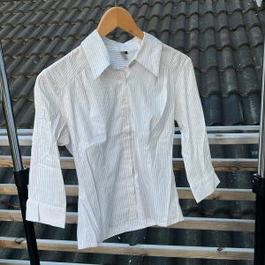 Vit randig skjorta från Vila - Vit skjorta från Vila med tunna grå ränder, klassisk krage och knäppning framtill. Skjortan har trekvartsärmar och figurnära passform. Perfekt för en clean och stilren look.
