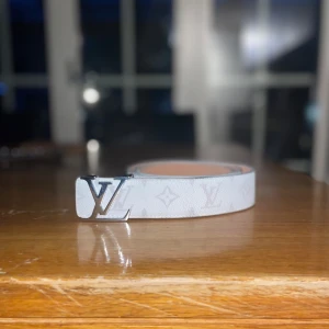 Vit Louis Vuitton monogram skärp - Snyggt vitt Louis Vuitton bälte i skinn med klassiskt monogrammönster och en silverfärgad LV-logga som spänne. Bältet har en stilren och modern look som verkligen sticker ut. Perfekt accessoar för att lyfta din outfit med exklusiv känsla.