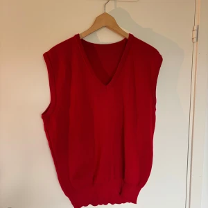 Röd  väst med v-ringning - En klassisk röd väst i tröjmaterial med v-ringad hals och ribbade kanter. Perfekt att slänga över en skjorta eller t-shirt för en snygg lager-på-lager-look. Västen är enkel och stilren utan mönster eller extra detaljer.