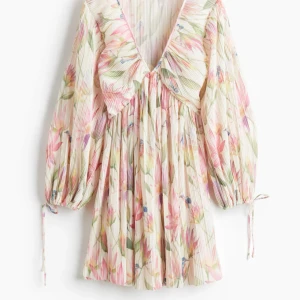 Blommig plisserad kortklänning - Superfin kortklänning med plisserat tyg och blommigt mönster i pastelliga toner av rosa, grönt och lila. Djup v-ringning, volangdetalj vid urringningen och långa puffärmar med knytband vid ärmslut. Perfekt för sommarens alla tillfällen!