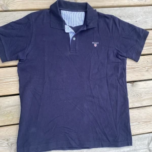 Marinblå pikétröja från GANT - Snygg marinblå pikétröja från GANT med klassisk krage och knäppning framtill. Tröjan har diskret logga broderad på bröstet och insida krage med blåvitt randigt tyg. Perfekt för en stilren och avslappnad look.